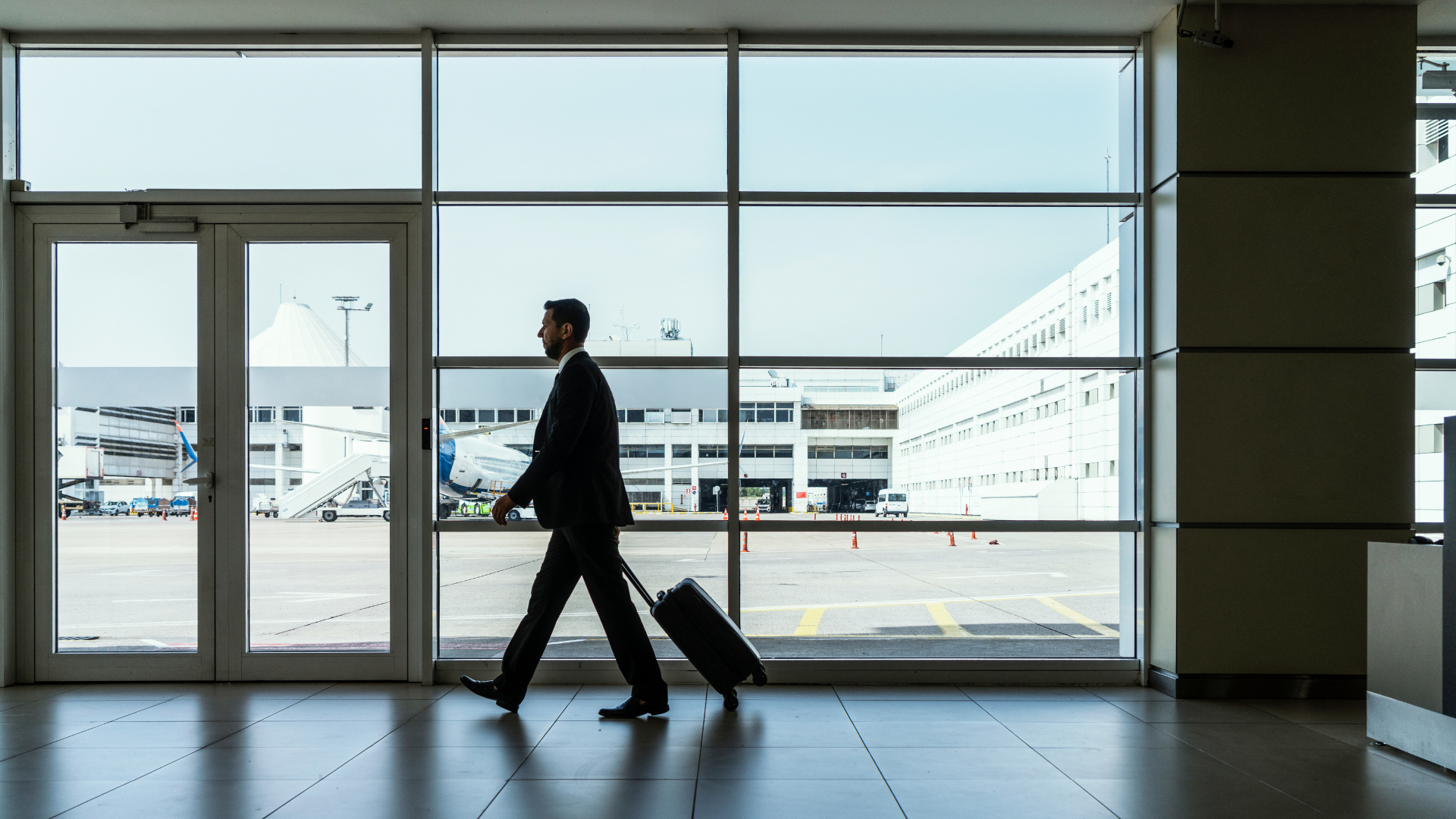 Business traveler walking through an airport terminal, pulling a rolling suitcase, with the runway and airport buildings visible through large windows.